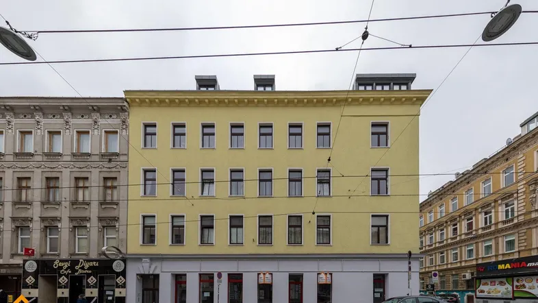 Ansicht eines mehrstöckigen Wohn- und Geschäftshauses mit gelber Fassade und vielen Fenstern an einer Straße.