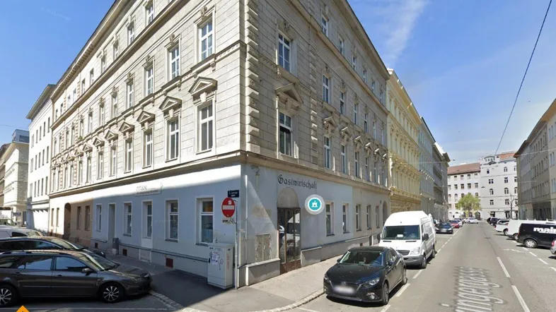 Mehrstöckiges Gebäude mit klassischer Fassade an einer Straßenecke in städtischer Umgebung, umgeben von parkenden Autos.