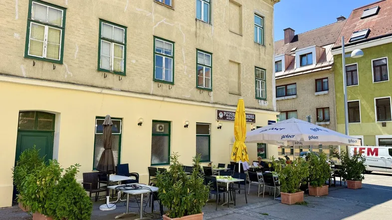 Außenansicht des Gebäudes mit einer belebten Restaurantterrasse und Sonnenschirmen an einem sonnigen Tag.