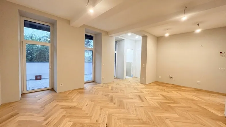 Geräumiger, heller Raum mit Fischgrätparkett und Zugang zu einem Außenbereich, ideal als Wohnzimmer.