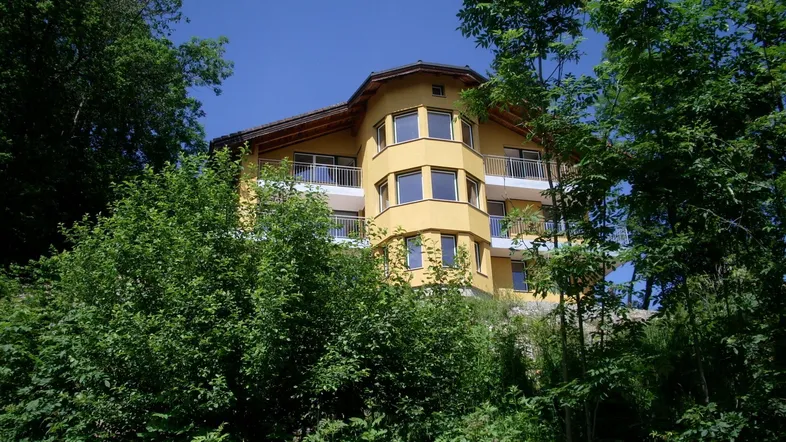 Gelbes Wohnhaus mit mehreren Balkonen, umgeben von dichtem Grün und hohen Bäumen.