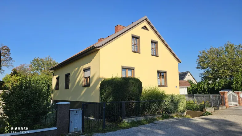 Gelbes Einfamilienhaus mit braunen Fensterrahmen und gepflegter Hecke unter blauem Himmel.