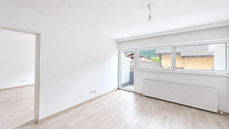 Helles, leeres Zimmer mit Laminatboden und Zugang zu einem kleinen Balkon.