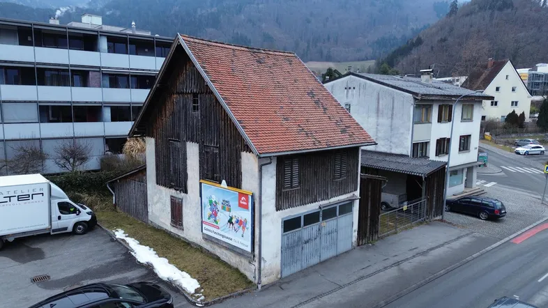 Altes Gebäude mit Holzelementen und Ziegeldach, umgeben von moderneren Bauten und einer Straße.