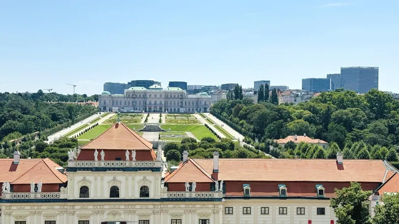 Panoramablick auf ein prächtiges historisches Schloss mit weitläufigen Gärten und moderner Stadtkulisse im Hintergrund.