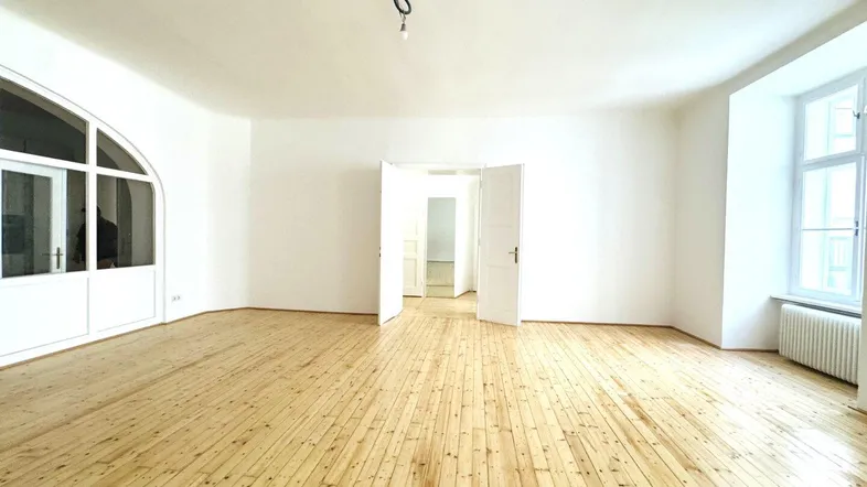 Helles, leeres Zimmer mit Holzdielenboden, weißen Wänden und großen Fenstern, bereit zur Einrichtung.