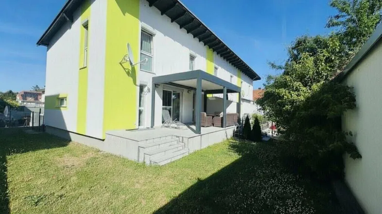 Modernes Einfamilienhaus mit farbiger Fassade, überdachter Terrasse und gepflegtem Gartenbereich.