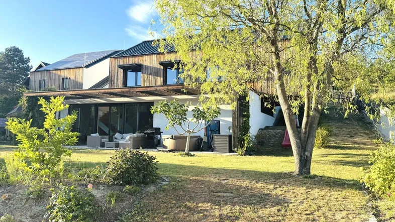 Haus mit Holzfassade, großer Terrasse und weitläufigem Garten unter blauem Himmel.