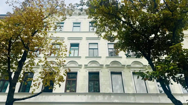 Historische Fassade eines Mehrfamilienhauses mit Stuckelementen und hohen Fenstern, umgeben von Bäumen.