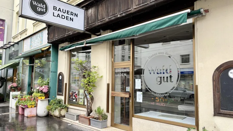 Ansicht der Ladenfront eines Bauernladens mit Schaufenstern und Markise an einem bewölkten Tag.