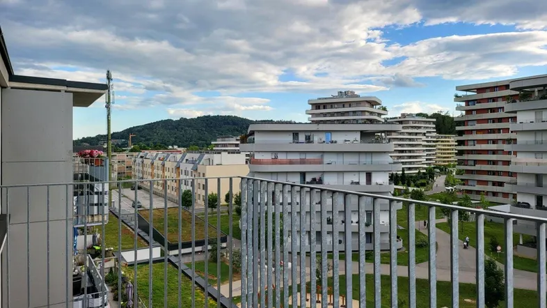 Blick vom Balkon auf moderne Wohngebäude, grüne Flächen und die umliegende Hügellandschaft.