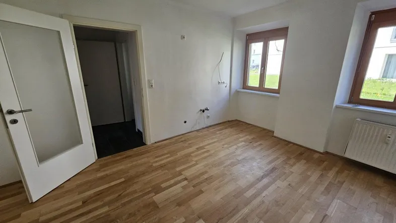 Leerer Raum mit Holzboden, zwei Fenstern und sichtbaren Anschlüssen an der Wand.