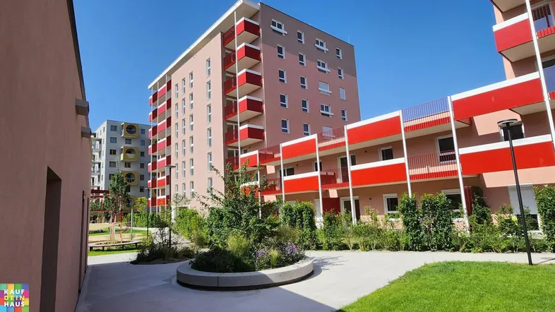 Moderne Wohngebäude mit roten Balkonen und gepflegten Grünflächen unter blauem Himmel.