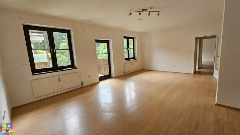 Geräumiges, leeres Wohnzimmer mit Laminatboden, großen Fenstern und Zugang zu einem Balkon.