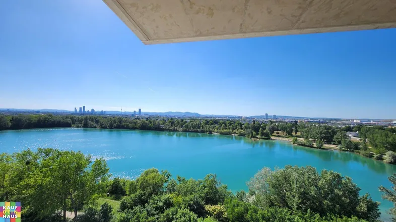 Panoramablick auf einen türkisfarbenen See mit grünen Ufern und der Skyline der Stadt im Hintergrund.