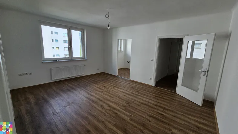 Helles, leeres Wohnzimmer mit Laminatboden und Blick auf ein gegenüberliegendes Gebäude.