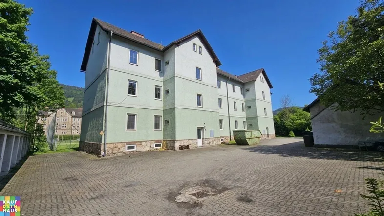 Mehrstöckiges Gebäude mit hellgrüner Fassade und Satteldach, umgeben von einem gepflasterten Hof und Bergen im Hintergrund.