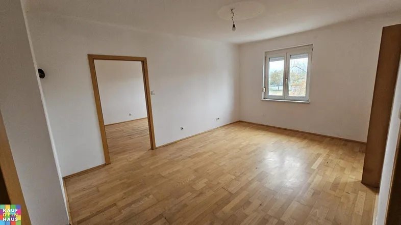Helles, leeres Zimmer mit Parkettboden und einem Fenster, das den Blick nach draußen freigibt.