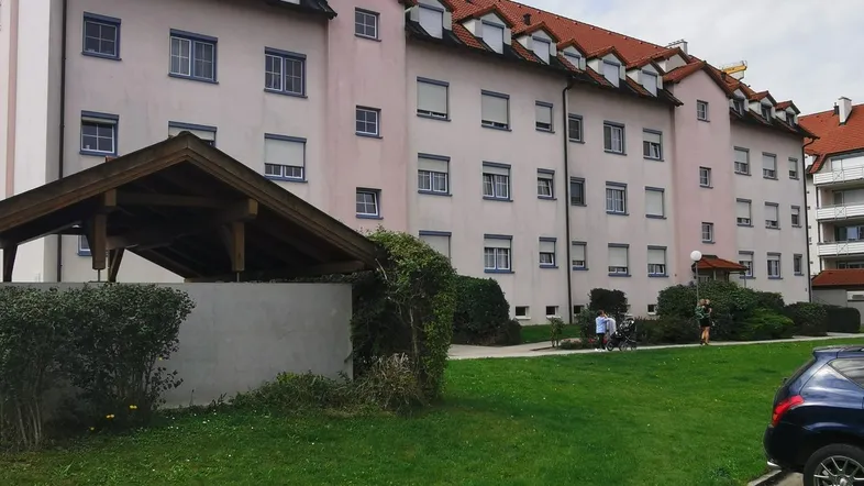 Mehrfamilienhaus mit heller Fassade, rotem Mansarddach und grüner Rasenfläche im Vordergrund.