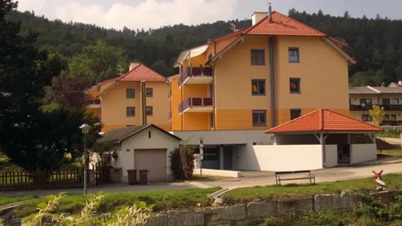 Mehrfamilienhaus mit gelber Fassade, roten Ziegeldächern und Balkonen in grüner Umgebung.