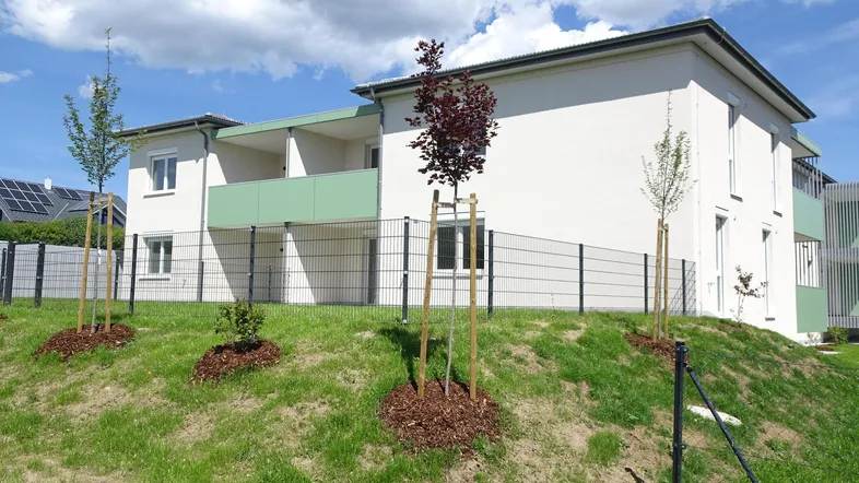 Neubaugebäude mit weißer Fassade, grünen Balkonen und jungen Bäumen in der Landschaft.