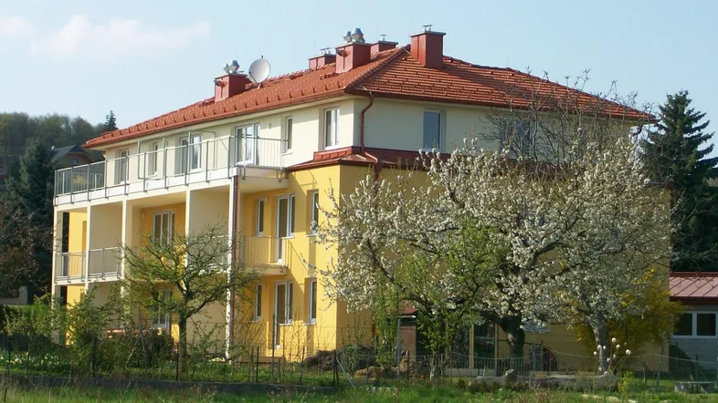 Mehrfamilienhaus mit gelber Fassade, roten Ziegeldächern und Balkonen in ländlicher Umgebung.