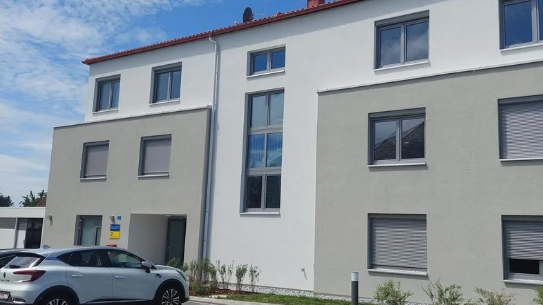 Modernes Mehrfamilienhaus mit heller Fassade, großen Fenstern und Außenparkplätzen unter blauem Himmel.