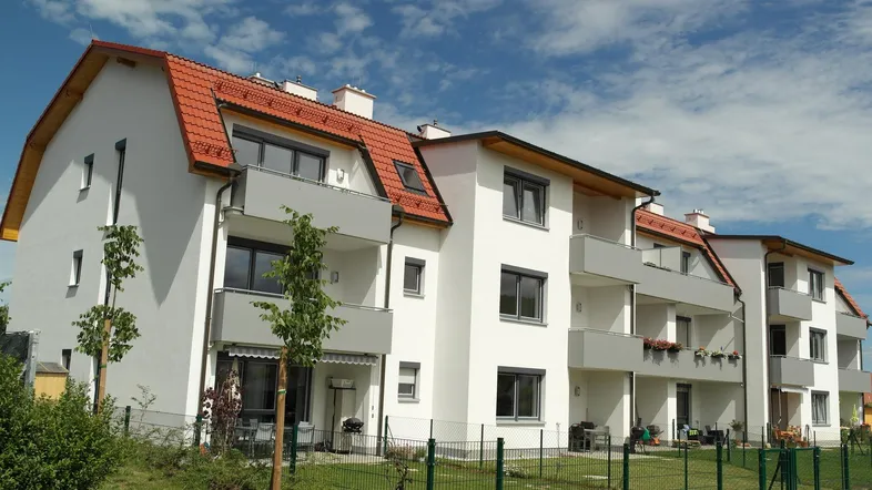 Modernes Mehrfamilienhaus mit weißer Fassade, roten Ziegeldächern und mehreren Balkonen unter blauem Himmel.