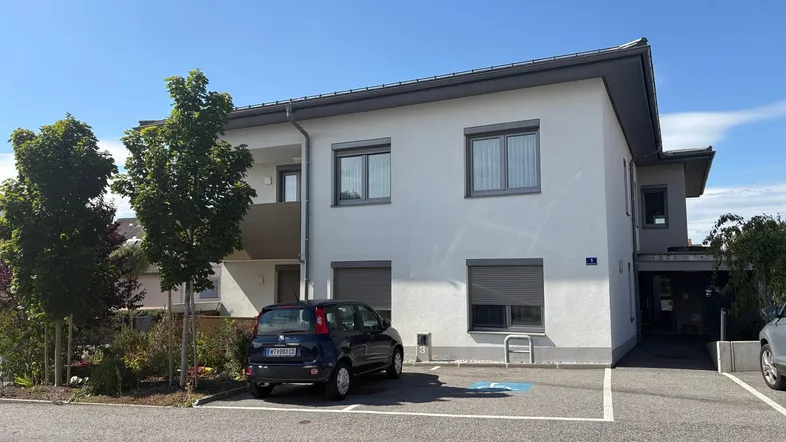 Modernes Mehrfamilienhaus mit heller Fassade, Balkon und Außenparkplätzen unter blauem Himmel.