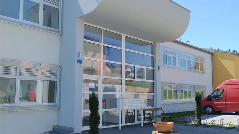 Modernes Gebäude mit großem Glaseingang und heller Fassade unter blauem Himmel.