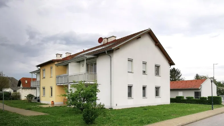 Mehrfamilienhaus mit weißer und gelber Fassade, Balkonen und gepflegtem Vorgarten unter bewölktem Himmel.