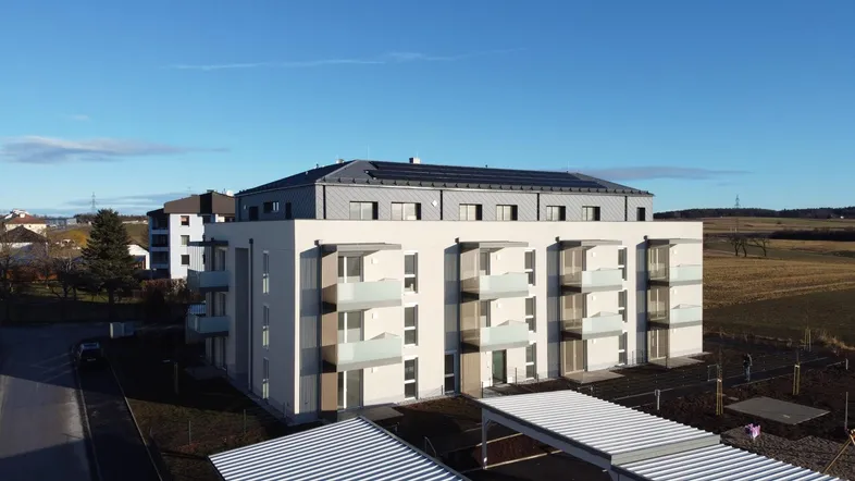Modernes Mehrfamilienhaus mit Balkonen und Photovoltaikanlage auf dem Dach, umgeben von ländlicher Landschaft.