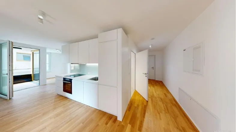 Heller offener Wohnbereich mit integrierter weißer Küchenzeile und Zugang zu einem weiteren Zimmer.