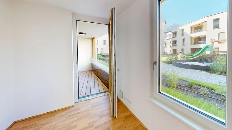 Heller Raum mit Holzboden und Zugang zu einem Balkon, Blick auf den Innenhof und Spielplatz.