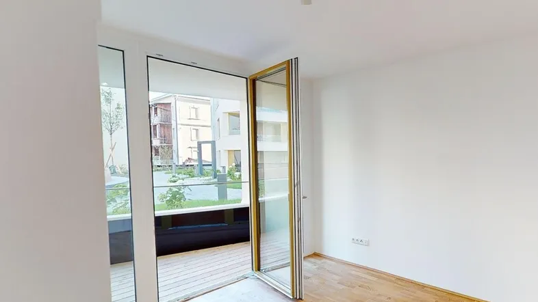 Heller, unmöblierter Raum mit Holzboden und großen Fenstern, die Zugang zu einem Außenbereich bieten.
