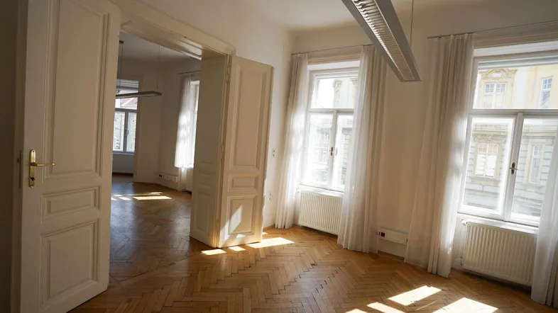 Heller, leerer Raum mit Fischgrätparkett, hohen Fenstern und viel Tageslicht, der in weitere Zimmer führt.