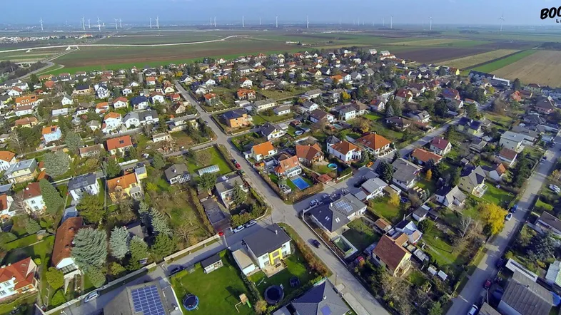 Luftaufnahme einer Wohnsiedlung mit Einfamilienhäusern, umgeben von Feldern und Windrädern in der Ferne.