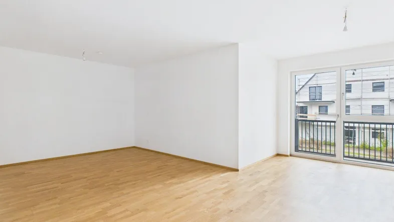 Helles, leeres Zimmer mit Parkettboden und großem Fenster, das Zugang zu einem Balkon bietet.