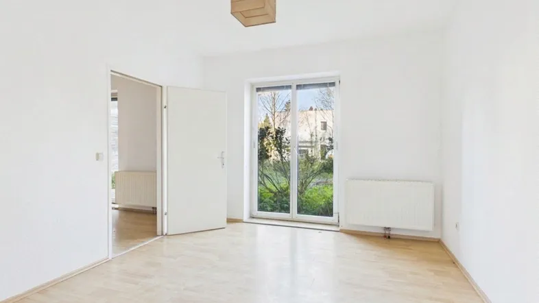 Helles, leeres Zimmer mit Parkettboden, weißen Wänden und Zugang zu einem grünen Außenbereich.