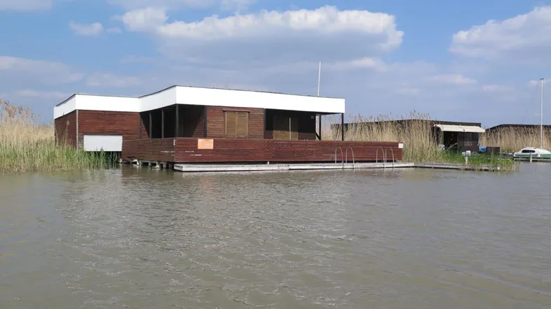 Hausboot auf dem Wasser mit Holzfassade und umlaufender Holzterrasse unter blauem Himmel.
