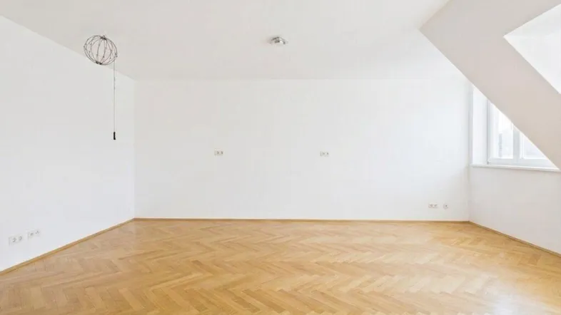 Helles, leeres Zimmer mit Fischgrätparkett und Dachschrägenfenster, bereit zur individuellen Gestaltung.