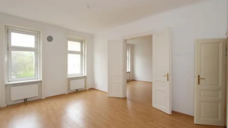 Heller, leerer Raum mit sauberem Laminatboden, zwei Fenstern und Zugang zu weiteren Zimmern.