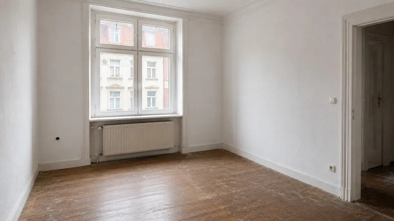 Heller Raum mit abgenutztem Holzboden, Stuckdecke und Blick auf die gegenüberliegende Fassade.