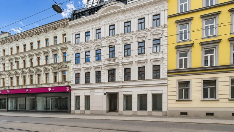Straßenansicht eines historischen Gebäudes mit reich verzierter weißer Fassade und Dachausbau, umgeben von weiteren Gebäuden.