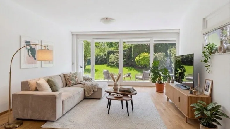 Helles Wohnzimmer mit beigem Sofa, Holzboden und großen Fenstern mit Blick in den Garten.