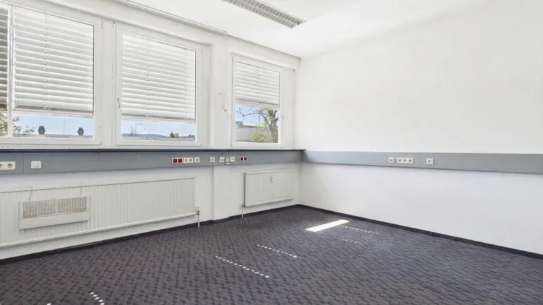 Helles, leeres Büro mit Teppichboden, großen Fenstern und Heizkörpern unter der Fensterbank.