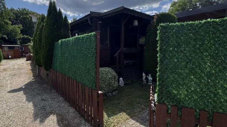 Kleines Holzhaus mit Kiesweg und grüner Hecke unter blauem Himmel an einem sonnigen Tag.