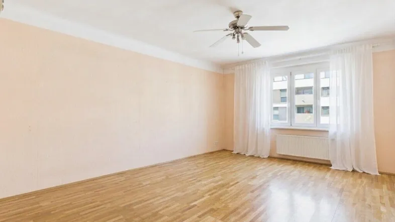 Helles, leeres Zimmer mit Parkettboden, Deckenventilator und großen Fenstern.