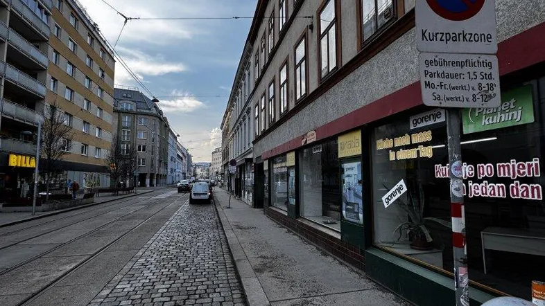 Blick auf eine belebte Stadtstraße mit Kopfsteinpflaster, historischen Gebäuden und Geschäften im Erdgeschoss unter blauem Himmel.