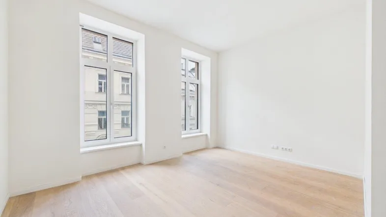 Helles, leeres Zimmer mit Holzboden und zwei großen Fenstern, die viel Tageslicht hereinlassen.